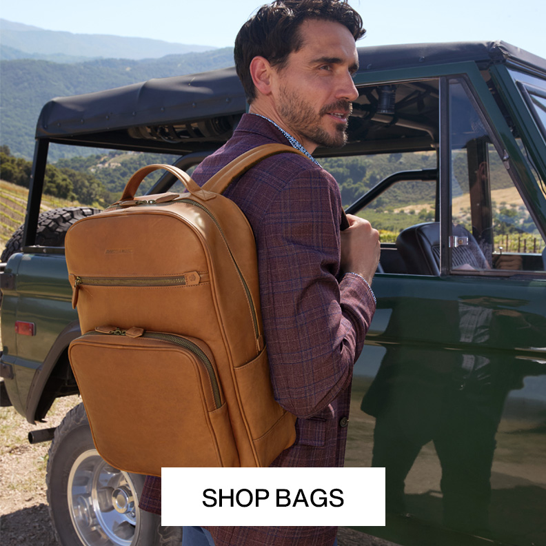 Man holding a tan leather backpack over one shoulder. He is standing outside in front of an old green colored jeep.