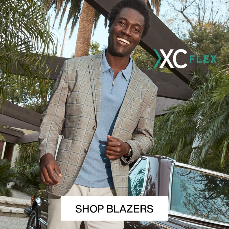 A man is pictured outside with tropical greenery surrounding him. He is wearing a gray plaid blazer, a light blue button-down shirt, and white pants with a Shop Blazers button at the bottom of the image.