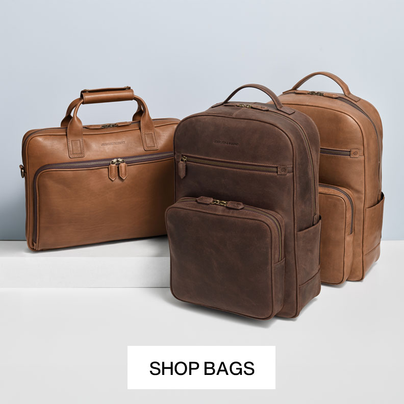 Man leaning on a vintage car holding onto a brown leather backpack with one hand. We see his blue colored pants and brown loafers.