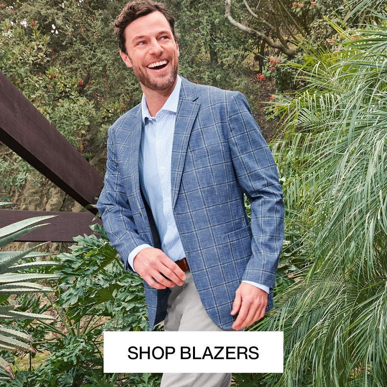 A man is pictured outside with tropical greenery surrounding him. He is wearing a blue plaid blazer, a light blue button-down shirt, and light grey pants.