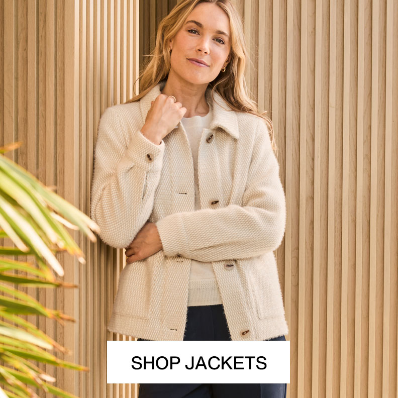 Woman wearing a cream textured jacket over a dark top, standing against a neutral colored wall with text that reads shop jackets.